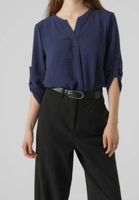 Blouse bleu marine à manches courtes, avec un col en V et des manches roulées, associée à un pantalon noir taille haute et une ceinture noire avec une boucle en argent.
