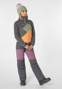 Giacca in pile con motivi geometrici in grigio, verde, arancione e rosa; abbinata a pantaloni grigi con accenti viola. Berretto lavorato a maglia verde.