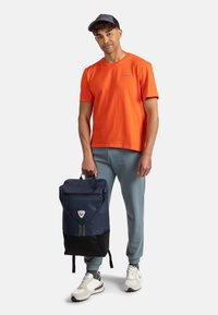 Zaino blu navy con una tasca frontale con zip, appoggiato su una base nera; indossato da una persona in una maglietta arancione e pantaloni della tuta azzurri chiari.