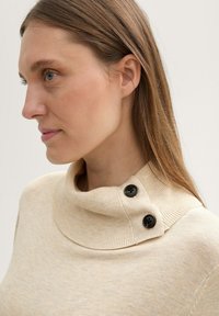 Beżowy sweter z dzianiny z kołnierzem typu funnel, ozdobiony dwoma czarnymi guzikami. Gładka tekstura, lekko luźny krój oraz szczegóły ribbingowe przy kołnierzu.