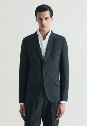 Joven con el cabello peinado hacia atrás, vestido con una chaqueta de traje oscura sobre una camisa blanca, posando frente a un fondo claro y liso.