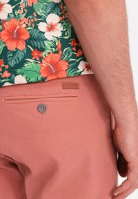 Kurzärmliges Blumenhemd mit mehrfarbigem Hibiskusdesign, kombiniert mit rosa Hosen mit einer Gesäßtasche und einem dekorativen Knopf.