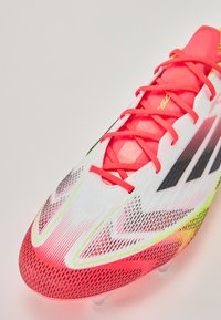 White and pink soccer cleat with red laces, featuring a textured upper, dynamic black and yellow accents, and a streamlined design.