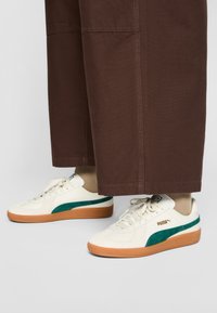Sneakers bianchi e verdi con un design testurizzato, caratterizzati da un logo dorato e una suola in gomma. Abbinati a pantaloni marroni a gamba larga.