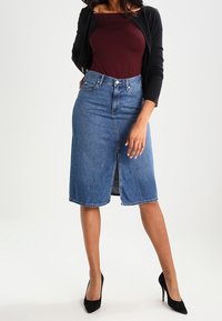 Falda de mezclilla de corte midi, color azul claro, con abertura frontal y diseño clásico de cinco bolsillos. Combinada con un cárdigan negro ligero.