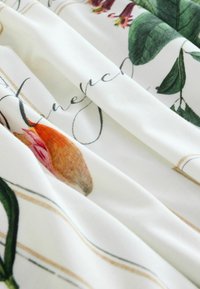 Valge kangas botaanilise mustriga, millel on rohelised lehed, oranžid ja roosad lilled ning mustas kursiivkirjas tekst pehmel volditud materjalil.