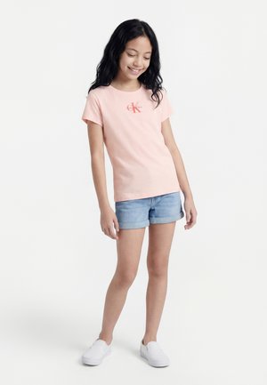 Κορίτσι που χαμογελά, φοράει ένα ανοιχτό ροζ T-shirt Calvin Klein, ανοιχτό μπλε τζιν σορτς και λευκά παπούτσια slip-on, στέκεται μπροστά σε ένα απλό φόντο.