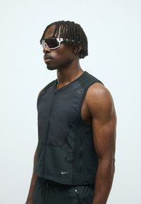 Schwarze Sportweste mit Reißverschluss vorne, strukturiertem Stoff und reflektierenden Details. Die Trägerin trägt außerdem dunkle Sonnenbrillen und geflochtenes Haar.