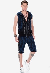 Marineblauer ärmelloser Hoodie mit einem strukturierten Muster, ausgestattet mit einem Frontreißverschluss und seitlichen Taschen, kombiniert mit passenden Shorts und weißen Turnschuhen.