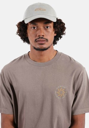 Homme aux cheveux bouclés portant une casquette beige clair et un t-shirt taupe avec un logo circulaire orange représentant un soleil et le texte "OXBOW" sur la poitrine.