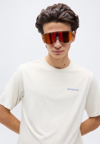 Νέος άνδρας που φοράει εκρού μπλουζάκι Patagonia και μεγάλα αθλητικά γυαλιά ηλίου με πορτοκαλί απόχρωση, ποζάροντας μπροστά σε απλό φωτεινό φόντο.