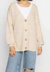 Beige pletený svetr s V-výstřihem, dlouhými rukávy a šesti velkými knoflíky. Textura je žebrovaná, v kombinaci s potrhanými světle modrými džíny.