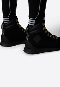 Bottes de randonnée en suède noir avec œillets en métal doré, semelle en caoutchouc et un patch noir marqué sur le talon, accompagnées de chaussettes noires.