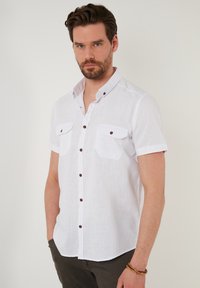 Camisa blanca de manga corta hecha de tejido texturizado, con dos bolsillos en el pecho, botones marrones y un diseño de cuello clásico.