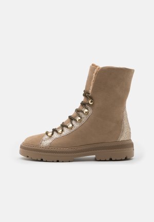 Taupefarbener, mittelhoher Wildlederstiefel mit dicker Sohle, goldenen Ösen, glitzerndem Stoffdetail, Fellfutter und Schnürung an der Vorderseite, seitlich fotografiert.