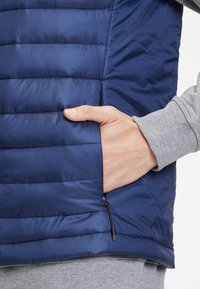 Marineblaues Steppweste mit horizontalem Steppmuster, ausgestattet mit einer seitlichen Reißverschlusstasche und einem grauen Pullover darunter.