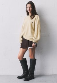 Pulover tricotat galben, fustă stratificată maro și cizme negre până la genunchi. Puloverul are o croială lejeră, cu textură vizibilă și un mic logo.