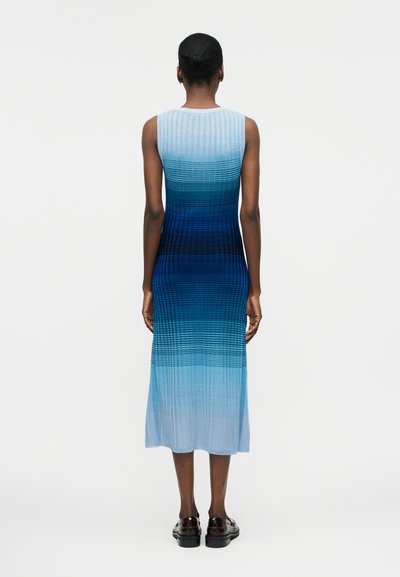 Γυναίκα που φοράει αμάνικο, ριπ πλεκτό φόρεμα με μπλε βαθμιδωτό χρώμα από ανοιχτό σε σκοτεινό, στέκεται πλάτη, με μαύρα παπούτσια.