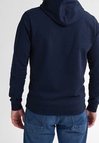 Tmavě modrá mikina s kapucí a dlouhými rukávy, s hladkou texturou a žebrovanými manžetami, kombinovaná s modrými džínami.