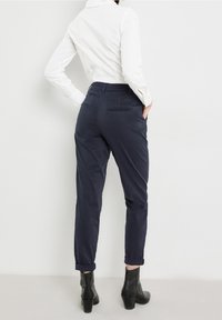 Navy trousers with a tapered fit, featuring rolled cuffs, side and back pockets, made of a smooth fabric. Worn with a white shirt.