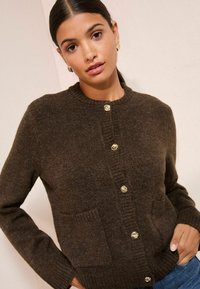 Brūns adīts džemperis ar apaļu kakla izgriezumu, zelta krāsas pogām, divām priekšējām kabatām un ribotiem piedurknēm un apakšmalu. Mīksta, teksturēta auduma.