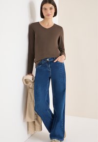 Braunes langärmeliges V-Ausschnitt-Oberteil kombiniert mit weitem blauen Jeans. Die Jeans haben einen Knopfverschluss und Fronttaschen, dazu ist eine beige Jacke über dem Arm gehängt.