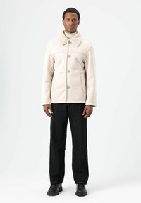 Lichtbeige jas met een schapenvachtkraag, knoopsluiting, zijzakken, gecombineerd met zwarte rechtgesneden broek en zwarte laarzen.