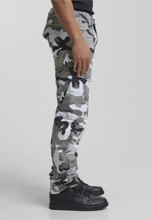 Hombre de pie con pantalones cargo de camuflaje en gris, blanco y negro combinados con zapatillas negras, sobre un fondo claro y liso.