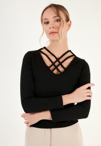 Maglia nera a maniche lunghe con costine, caratterizzata da una profonda scollatura a V con spalline incrociate. Tessuto liscio, design aderente e silhouette semplice.