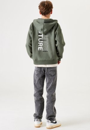 Olivově zelená mikina s kapucí s bílým nápisem na zadní straně, kombinovaná se šedými džíny a bílými teniskami s černými detaily. Hladká látka a volný střih.