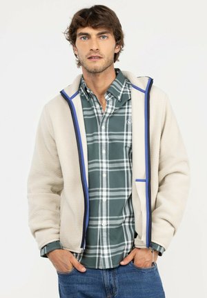 Hombre con cabello castaño que lleva una chaqueta de forro polar beige con cremallera sobre una camisa de cuadros verde y blanca y vaqueros azules, de pie con las manos en los bolsillos.