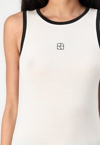 Valkoinen tank-top mustilla ribattuja reunuksilla, vartalon myötäinen muotoilu ja pieni musta logo edessä. Kangas näyttää sileältä ja joustavalta.