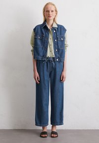 Denim vest met grote zakken, gecombineerd met een lichtgroen overhemd met knopen en een brede, blauwe denimbroek. Zwarte instapsandalen.