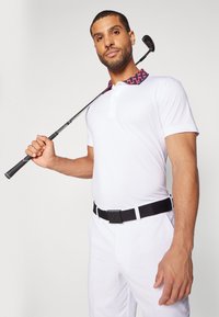 Λευκό πόλο πουκάμισο με σχέδιο στον γιακά, μαύρη ζώνη και λευκό παντελόνι. Το μοντέλο κρατάει ένα μπαστούνι γκολφ, αναδεικνύοντας έναν αθλητικό σχεδιασμό.