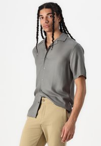Short-sleeve, grey shirt made of smooth fabric, featuring a collar and green buttons, paired with beige trousers.