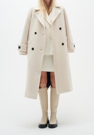 Mujer con un abrigo largo crema de doble botonadura, falda a juego y botas beige hasta la rodilla con suelas negras, sobre un fondo blanco.