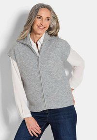 Hellgraue gestrickte ärmellose Weste mit hohem Kragen, Reißverschluss und geripptem Textil, getragen über einem weißen Hemd und kombiniert mit dunklen Jeans.