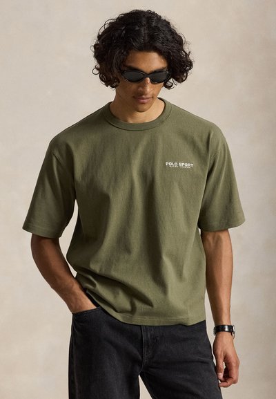 Άνδρας με σκούρα γυαλιά ηλίου, ελιάς "Polo Sport Ralph Lauren" κοντομάνικη μπλούζα, μαύρο παντελόνι και ρολόι χειρός, στέκεται μπροστά σε απλό φόντο.