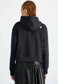 Čierna mikina s kapucňou z mäkkého materiálu, skráteného dizajnu, so sťahovacou šnúrkou a logom na chrbte. Doplnená elegantnou čiernou plisovanou sukňou.