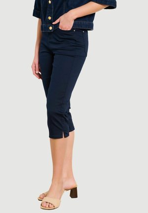 Naine, kes kannab tumesiniseid lühikesi pükse eesmistesse taskutesse käega toetades, seljas beežid kontsakingad, taust on lihtne.