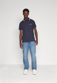 Polo shirt blu navy con colletto a righe bianche, maniche corte e logo anteriore, abbinato a jeans chiari strappati e sneaker bianche.