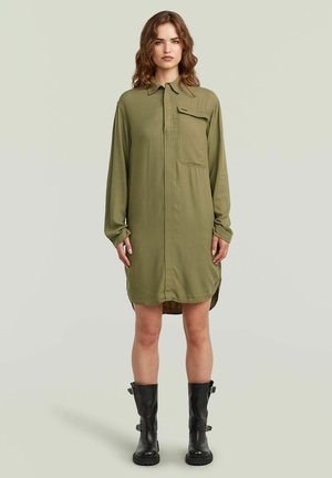 Frau mit welligem Haar, die ein olivgrünes Hemdkleid mit langen Ärmeln und schwarze Stiefel bis zur Mitte der Wade trägt, steht vor einem schlichten hellen Hintergrund.