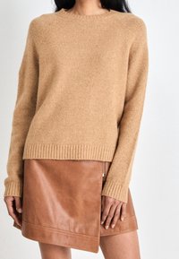 Kvinna iklädd en beige stickad tröja och en brun läderkjol med asymmetrisk design och sidogylf, stående mot en neutral bakgrund.