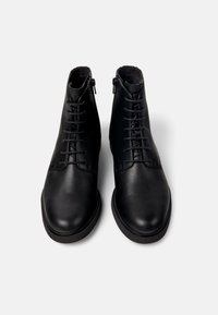 Botas de tobillo de cuero negro con una textura suave, que presentan un cierre de cordones en la parte frontal y un cierre lateral para facilitar el uso. Suela de goma plana.