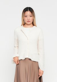 Junge Frau mit langen Ombre-Haaren, die eine weiße Strickjacke mit Knöpfen über einem weißen Oberteil und einem braunen Satrock trägt, vor einem weißen Hintergrund steht.
