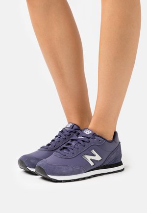 Jalat yang mengenakan sepatu New Balance ungu dengan sol putih dan hitam, ditampilkan di latar belakang putih polos.