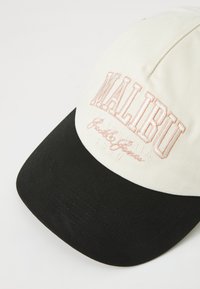 Pallikeer kaabu, millel on valge kangast kroon ja must visor. "MALIBU" on roosas värvuses tikandina esiosas, all on lisatekst.