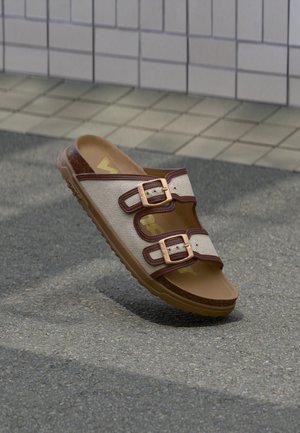 Sandalo beige con due fibbie regolabili marroni, sottopiede color cuoio e suola marrone texturizzata su pavimentazione grigia vicino a una parete piastrellata.