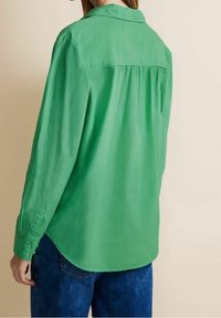 Grön skjorta med krage och plisserad bakdetalj, långa ärmar, knäppning vid manschetterna och en avslappnad passform. Tillverkad av lätt tyg med en mjuk textur.