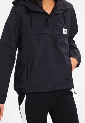 Person trägt eine schwarze Kapuzenjacke mit Frontreißverschluss und Logopatch, steht mit einer Hand in der Tasche, kombiniert mit schwarzen Hosen mit weißen Seitenstreifen.